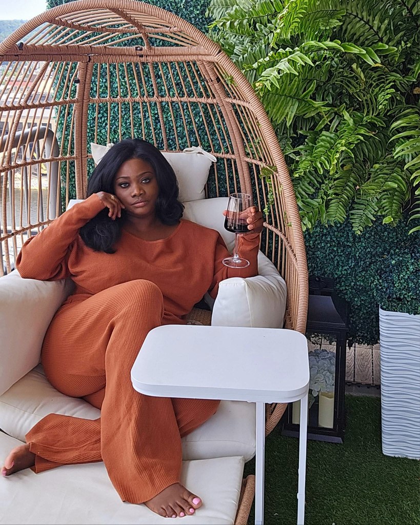 A Black woman relaxing outdoors in a rust-colored lounge set, holding a glass of wine, seated in a cozy egg chair surrounded by greenery symbolizing soft life, self-care, and emotional rest.