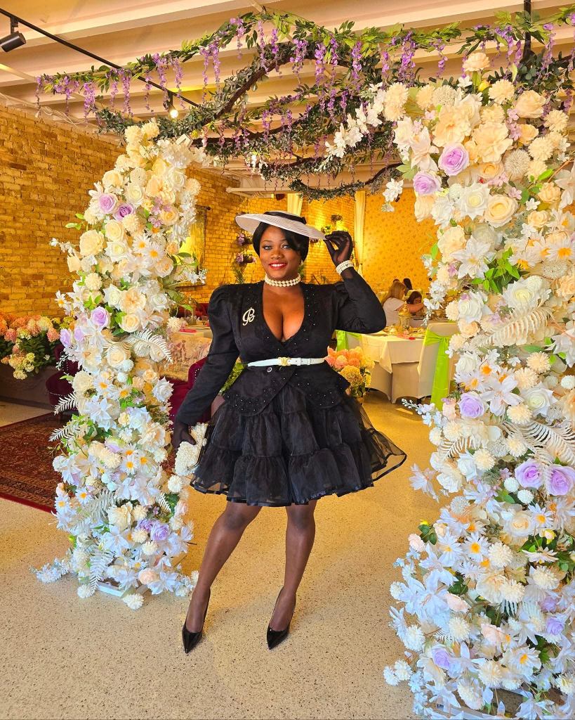 Elegant woman in a black ruffle dress and hat posing under a floral arch at a summer tea party