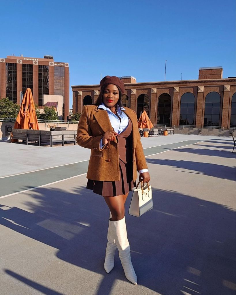 Preppy fall outfit with brown blazer, pleated mini skirt, blue button-down shirt, beret, cream knee-high boots, and structured handbag on a city rooftop.