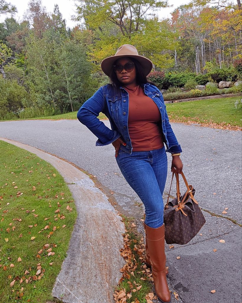 Fall casual outfit with denim jacket, rust top, skinny jeans, brown knee-high boots, wide-brim hat, and Louis Vuitton tote on a tree-lined street.