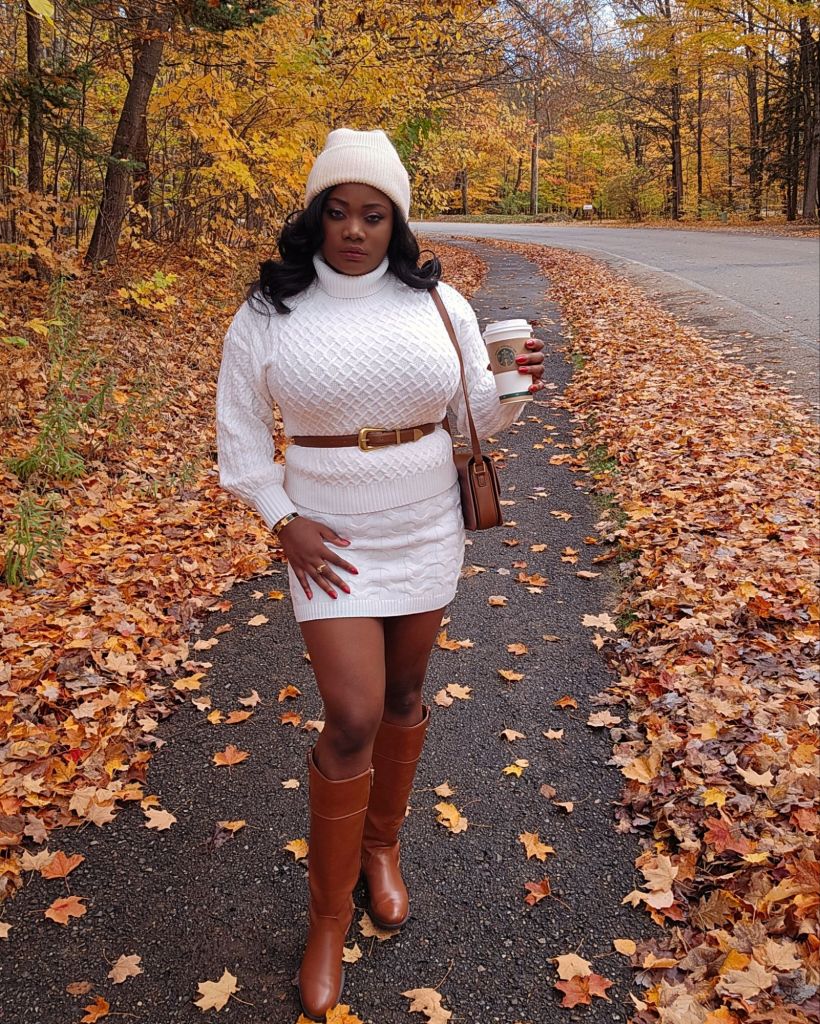 Beatrice Mwansa walking along a tree-lined path covered with autumn leaves, wearing a cozy white knit turtleneck sweater dress paired with a beige beanie, brown belt, brown leather crossbody bag, and knee-high tan boots. She holds a Starbucks coffee cup and smiles softly while touching her hat, surrounded by golden fall foliage.
